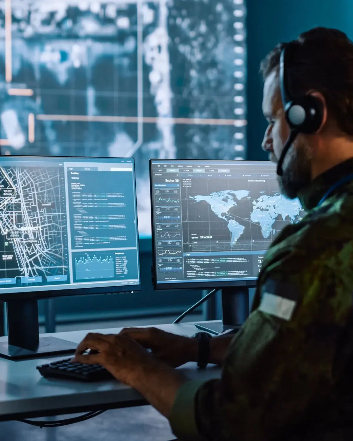 A military or security operator wearing a headset monitors multiple screens displaying world maps, data, and geographical information in a high-tech control room