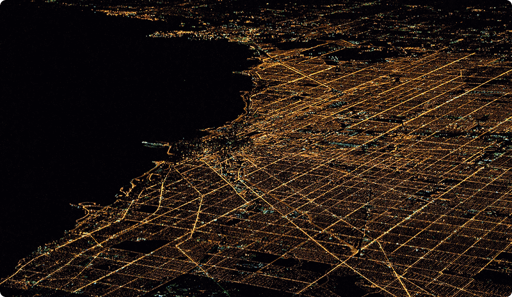 An aerial night view of a sprawling coastal city with illuminated streets forming a distinct grid pattern