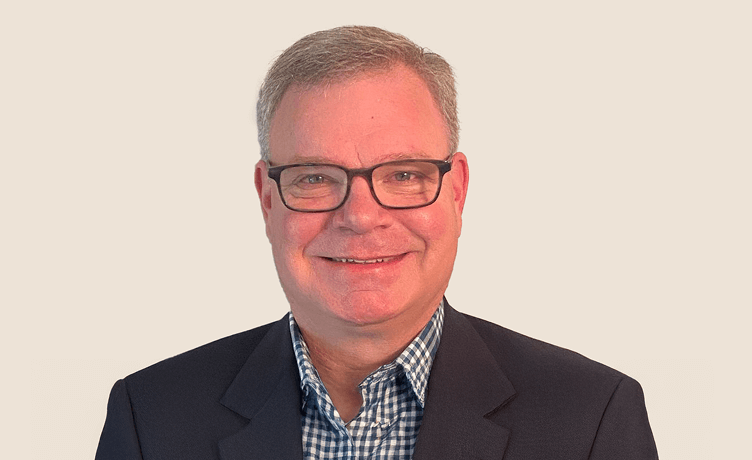 A professional headshot of a smiling middle-aged man wearing glasses and a dark suit jacket over a checkered shirt
