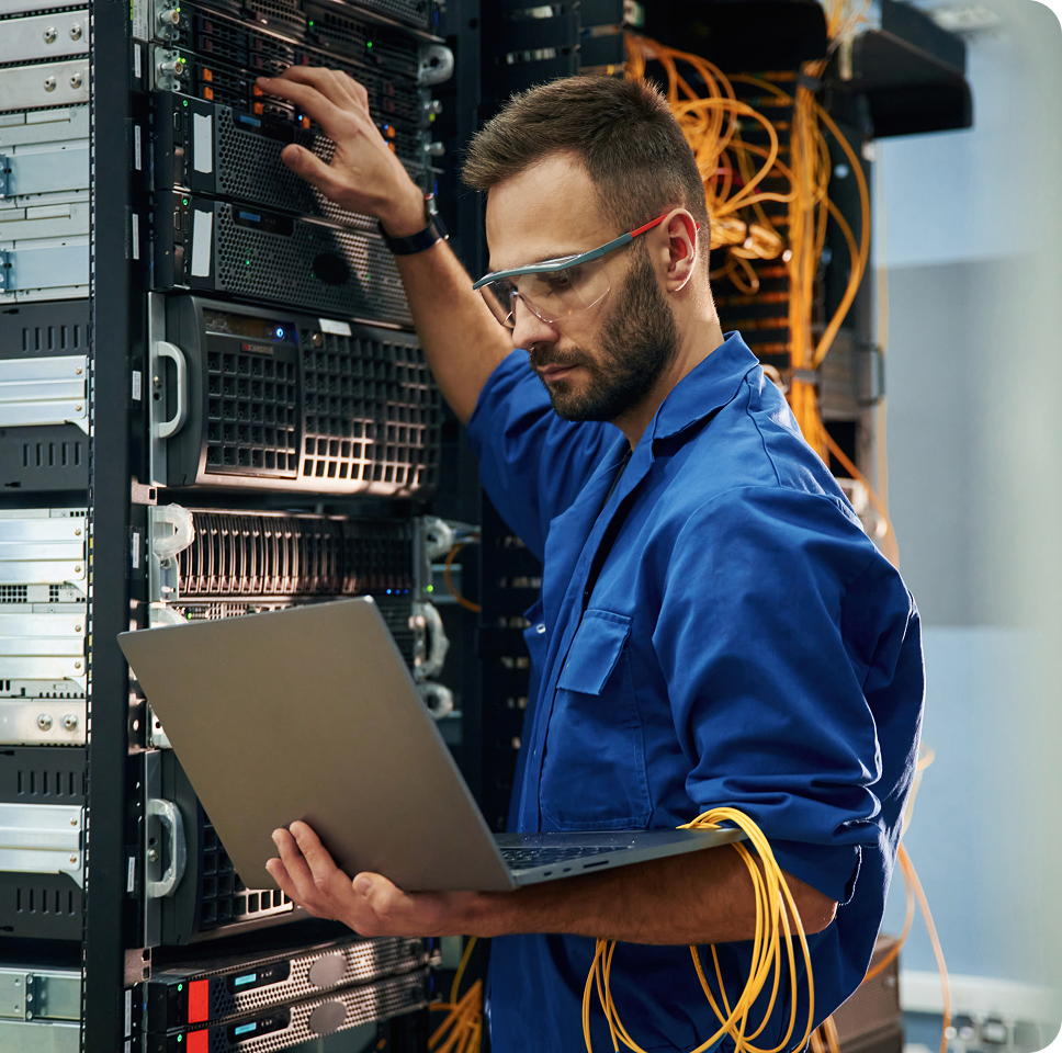 An IT technician in a blue uniform and safety glasses working in a server room with a laptop